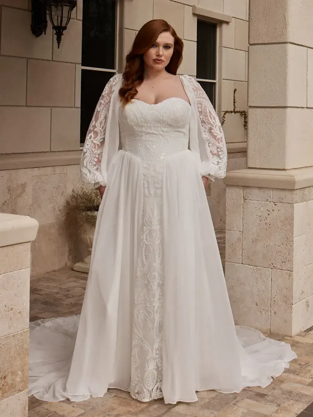 A woman in a white wedding dress with a lace bodice and sleeves, standing outside on a stone patio.