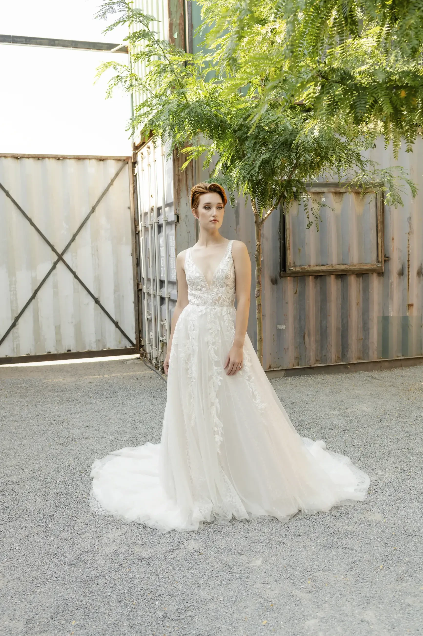 Bride in a flowing white gown with lace details stands in a sunlit, gravel courtyard. Rustic metal containers and lush greenery frame the serene scene.