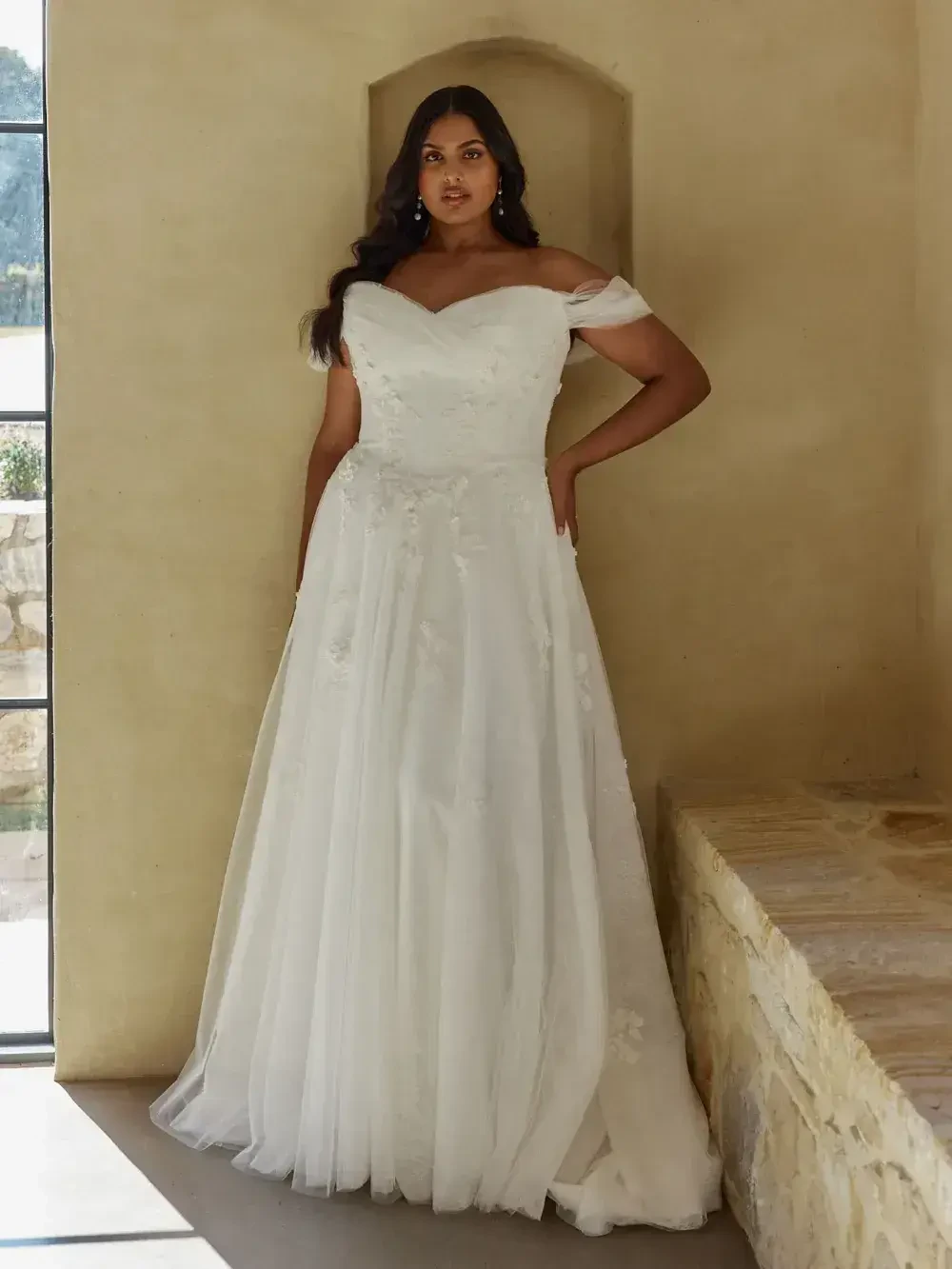 Bride wearing an elegant off-shoulder white wedding gown with floral details stands in a softly lit room, exuding confidence and grace.