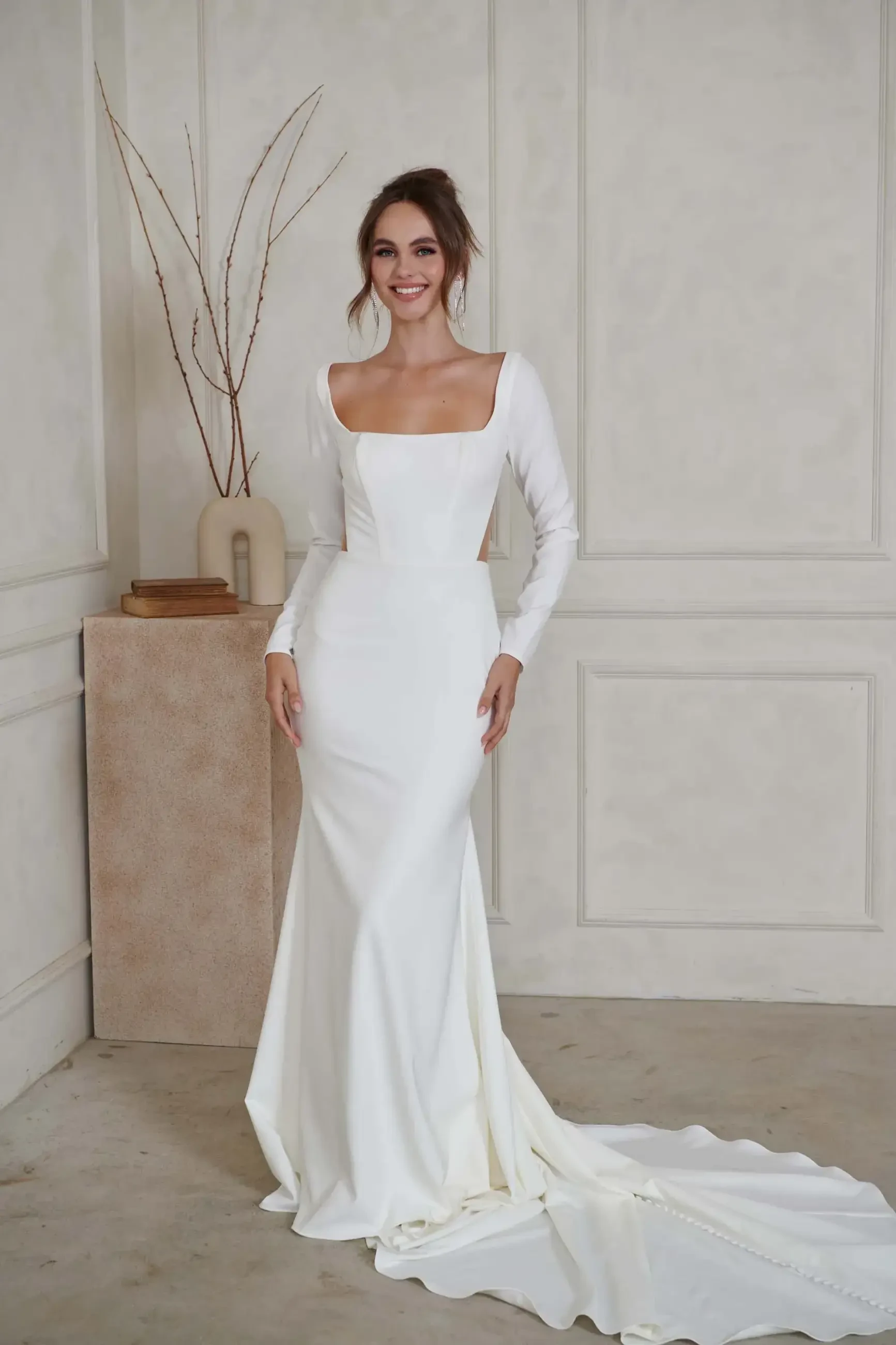 A woman in an elegant white wedding dress stands in a minimalist room, with a soft smile. The gown features long sleeves and a flowing train.