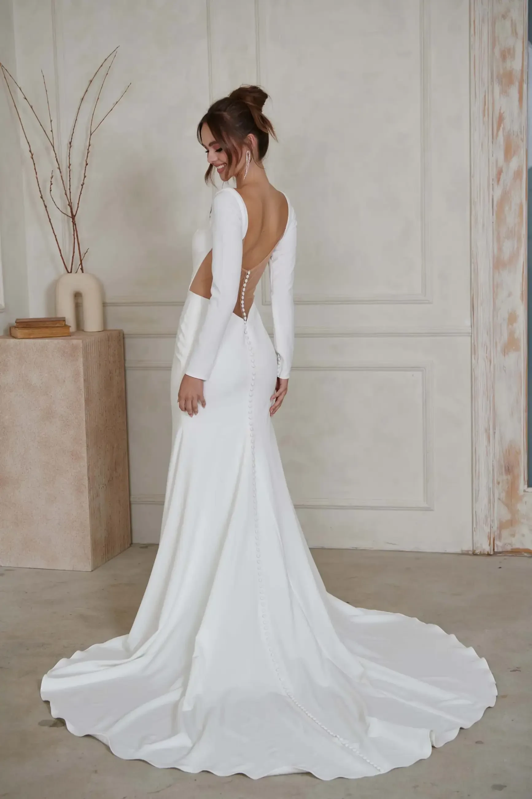 Bride in elegant long-sleeved white wedding gown with open back and button detail, stands in a minimalist room with neutral tones and modern decor.
