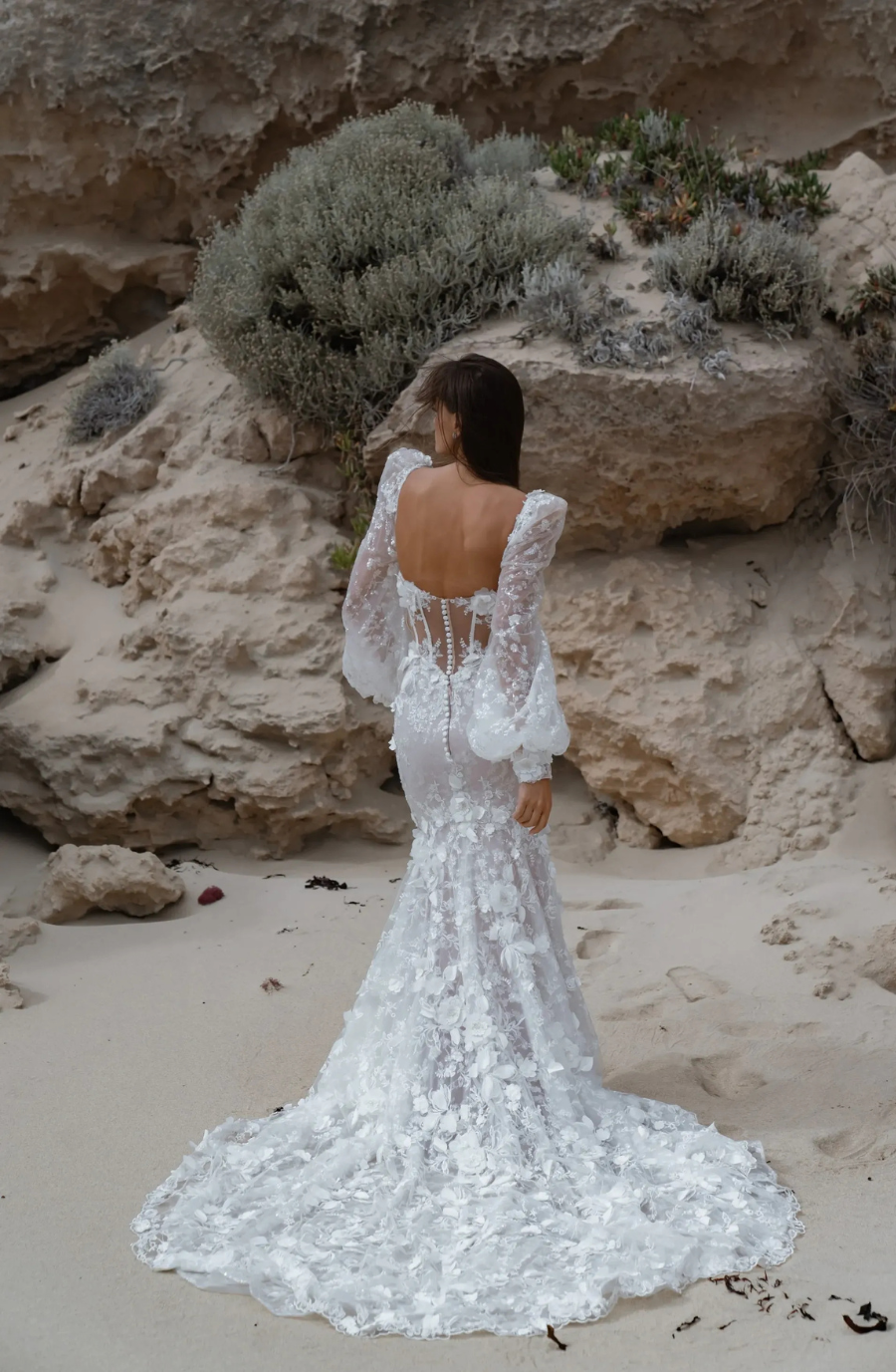 Bride in a lace wedding dress with a long train stands on a sandy beach, surrounded by rocky cliffs and greenery, conveying elegance and tranquility.