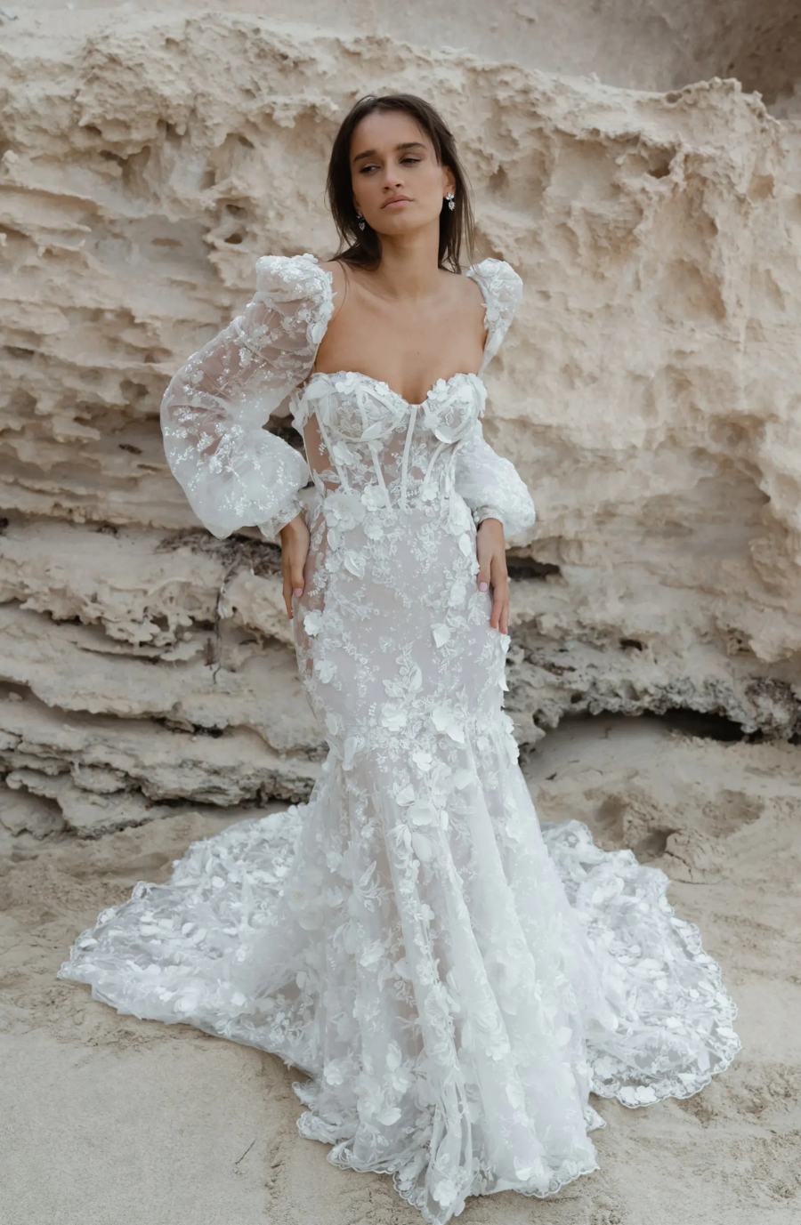 Woman in an elegant, lace bridal gown with floral patterns stands on a sandy beach. Her expression is serene, and the rocky background adds contrast.