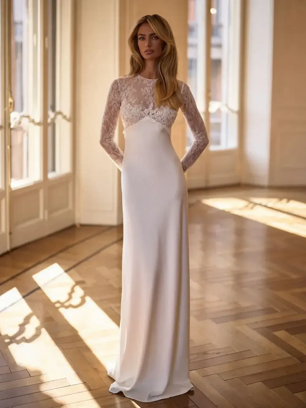 A woman stands in a bright room wearing a long, elegant white dress with lace details on the top. The dress has long sleeves and a sleek silhouette, and she is posed with her hands in the pockets of the gown.