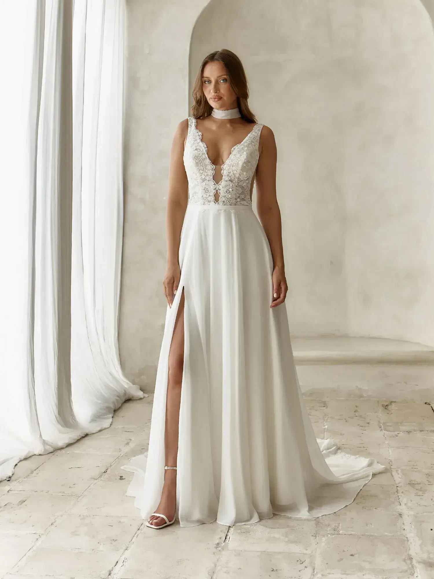 A model is wearing a white wedding dress with a plunging neckline and lace detailing. The dress has a thigh-high slit and flows down to the floor. The background features light-colored walls and draped fabric.