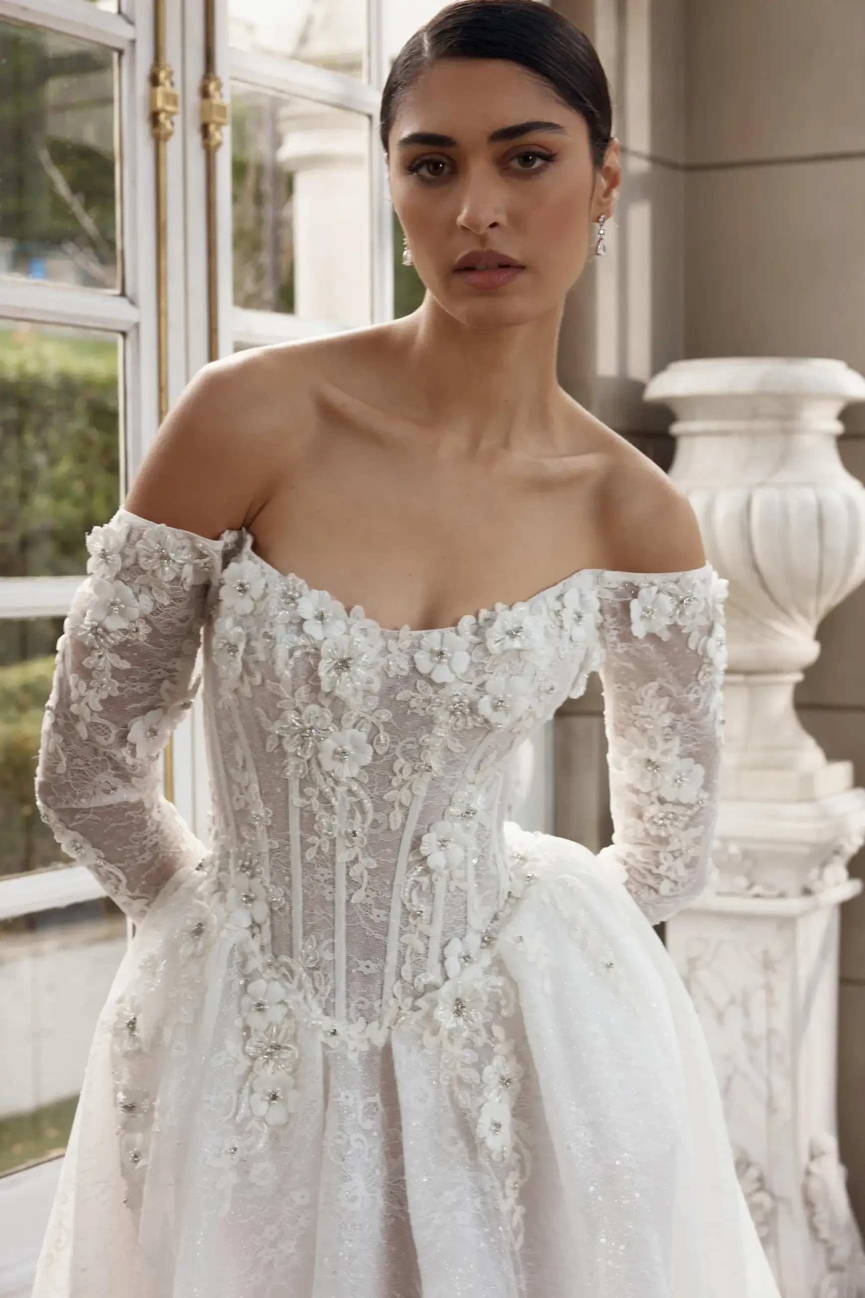 A model wearing an off-the-shoulder wedding dress adorned with floral embellishments and long lace sleeves, posing in a well-lit room with large windows.