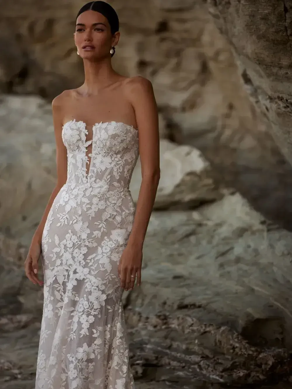 A model stands elegantly in a strapless, intricately designed white lace wedding dress against a rocky background.