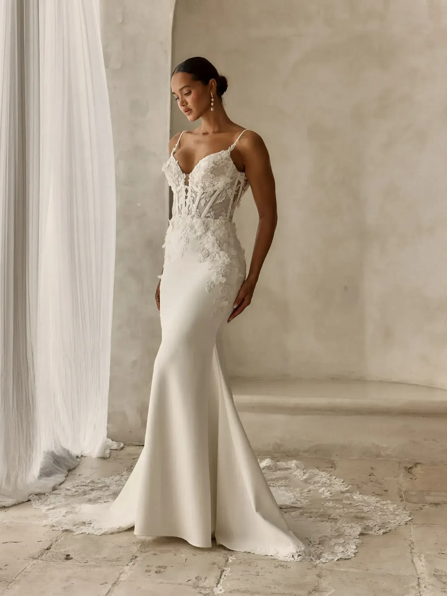 A woman wearing an elegant white wedding dress with intricate lace detailing, standing gracefully against a neutral background.