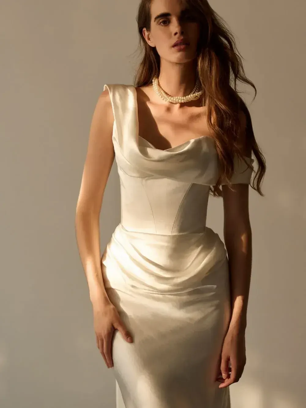 A model wearing a elegant, fitted white dress with a cowl neckline, styled with a pearl necklace and soft waves in her hair.