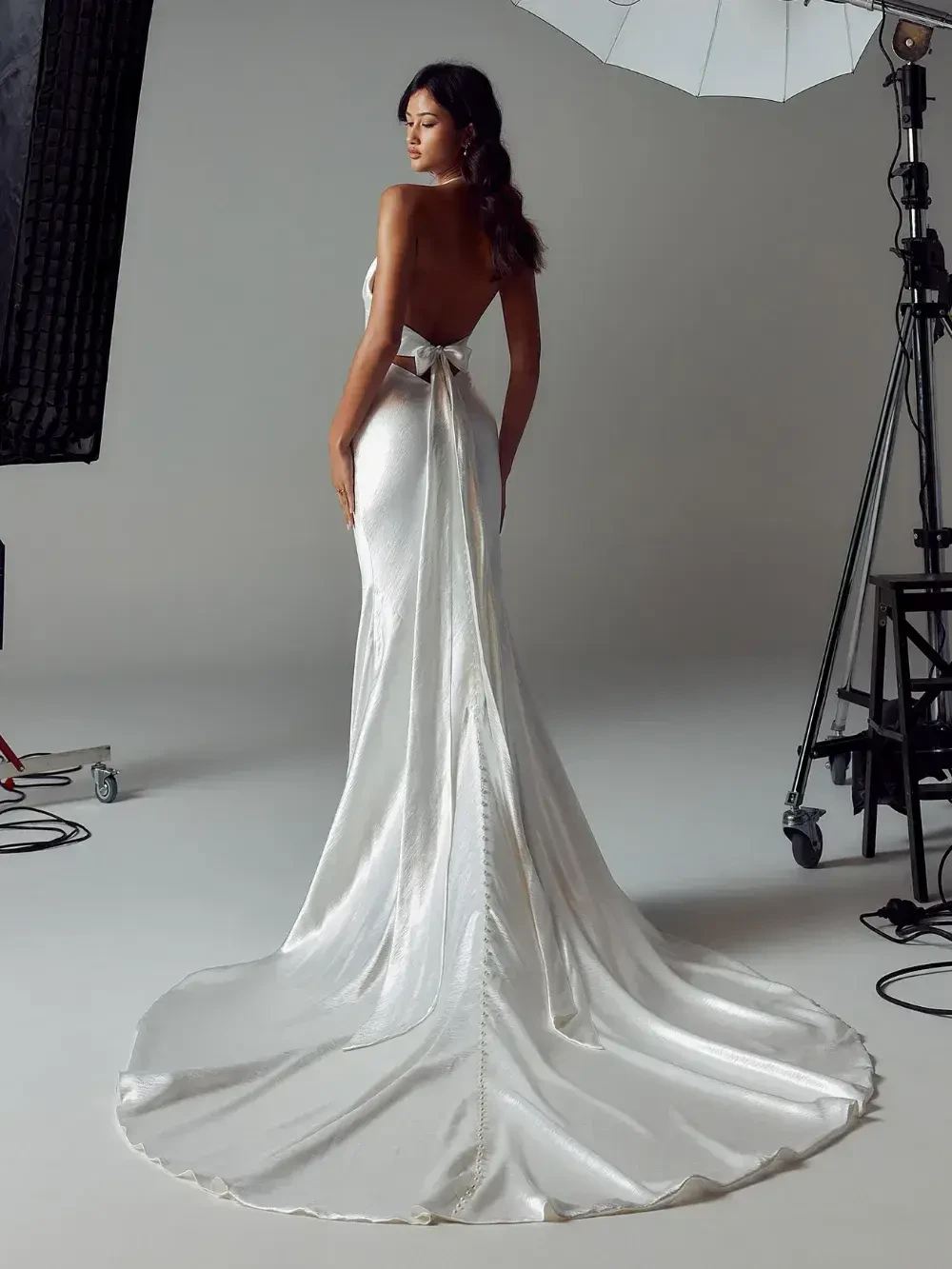 A model wearing a backless white gown with a train, posed in a photo studio setting with professional lighting equipment.