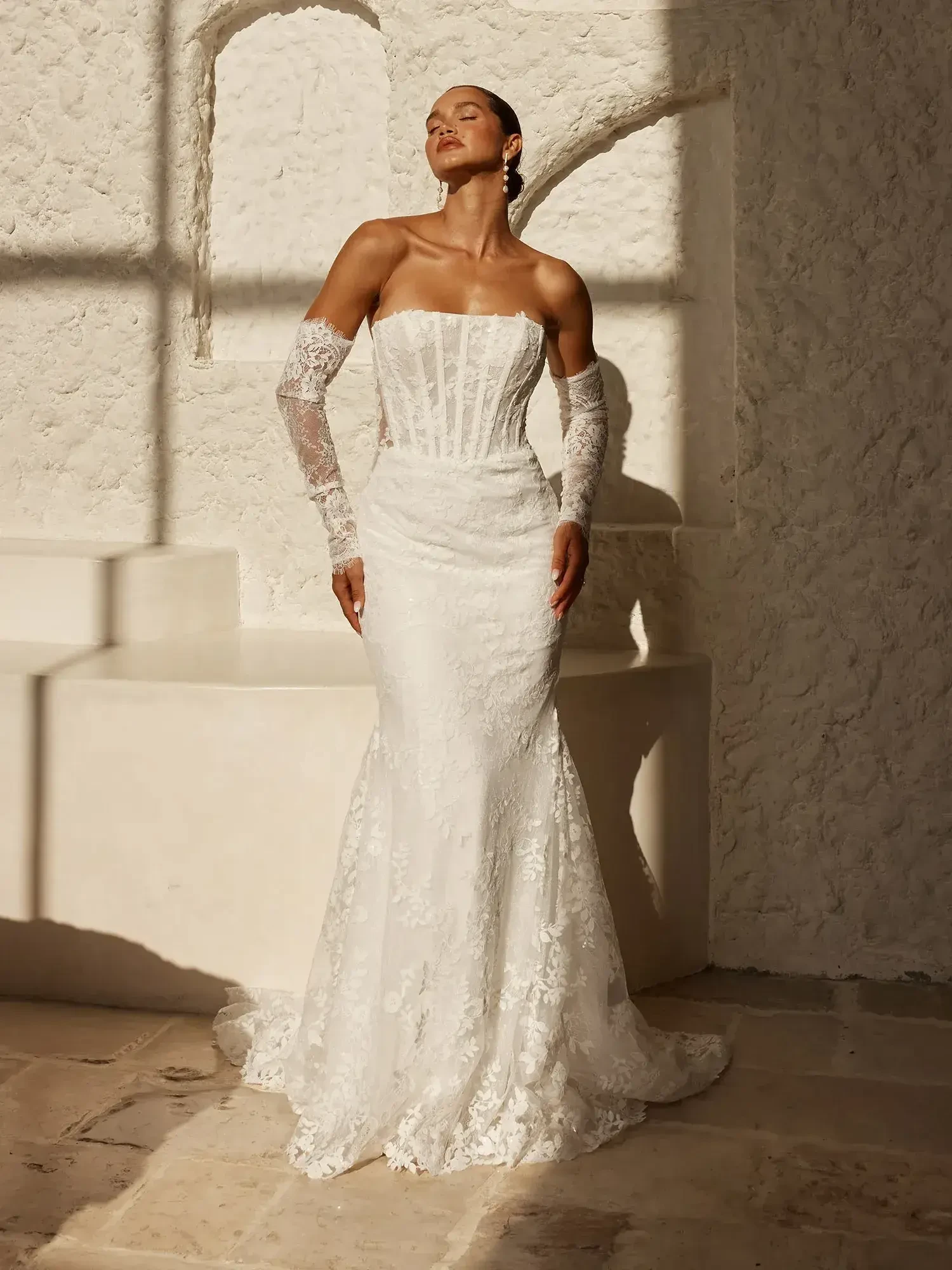 A model poses in a fitted white wedding dress with lace details and strapless design, featuring long sheer sleeves, standing in a sunlit room with textured walls.